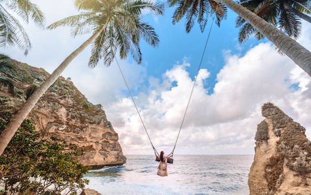 Person on a swing between palm trees overlooking ocean cliffs, Nusa Penida Island.