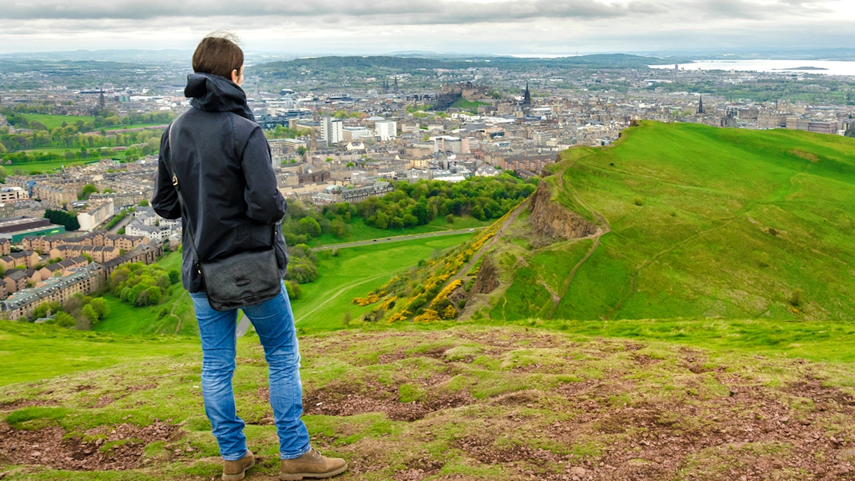 Person overlooking Edinburgh cityscape from a hilltop, with Edinburgh Castle visible in the distance.