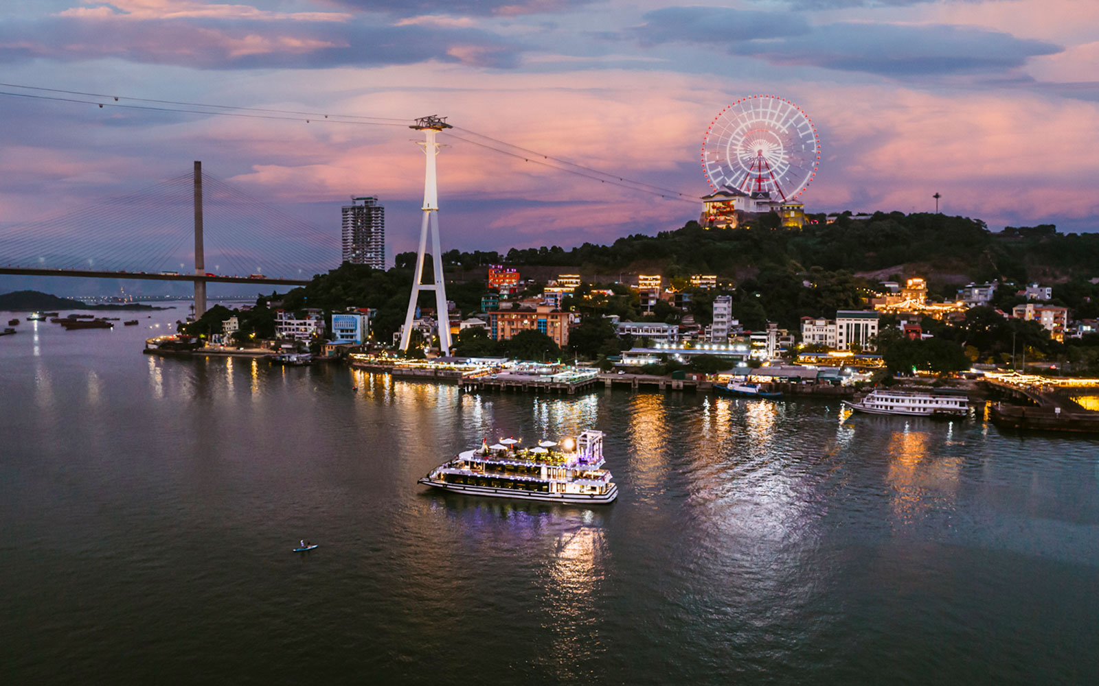 Cruise ship on Ha Long Bay at sunset with city lights and Ferris wheel in the background.