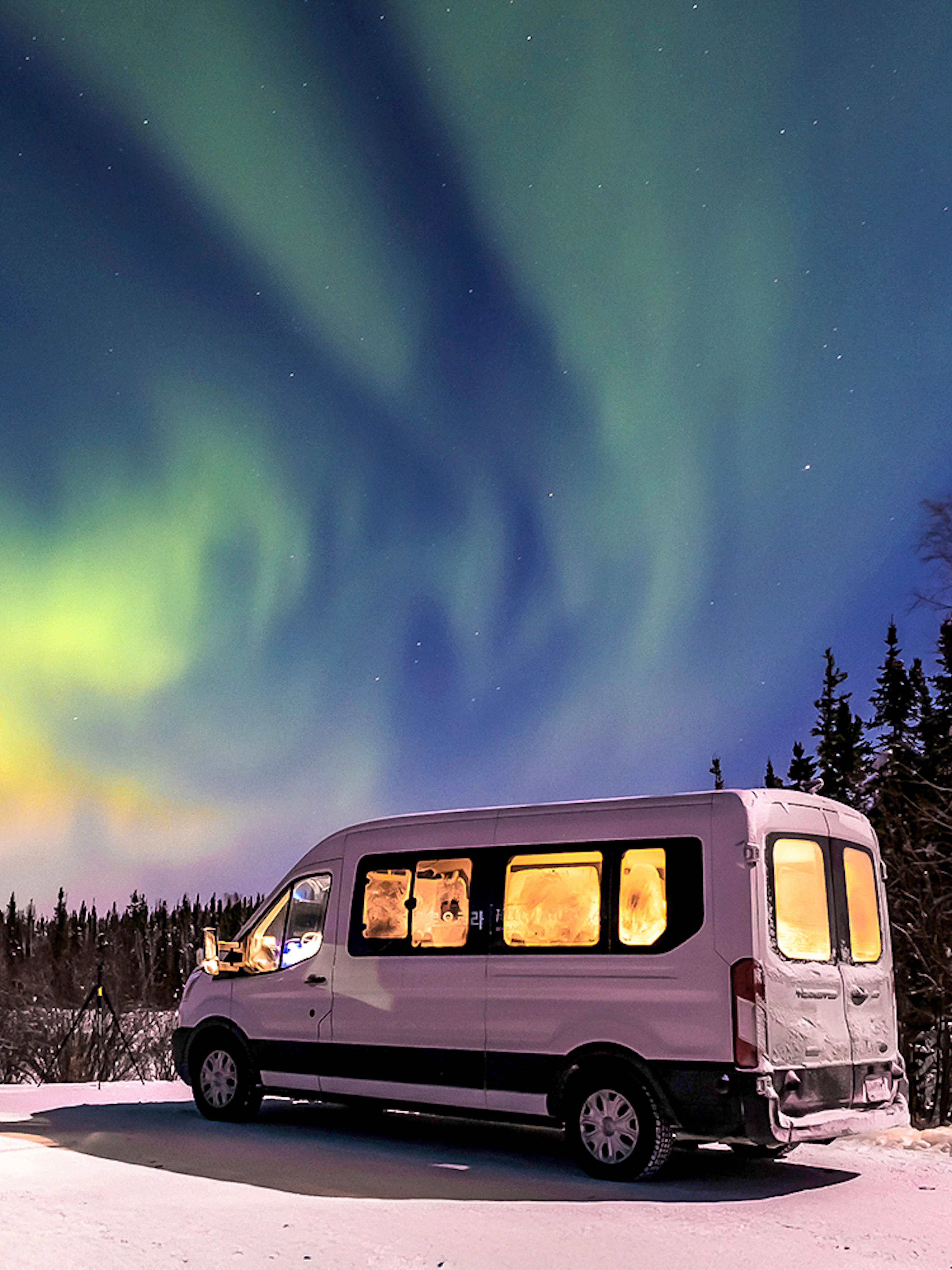 Minibus under Northern Lights in snowy landscape.