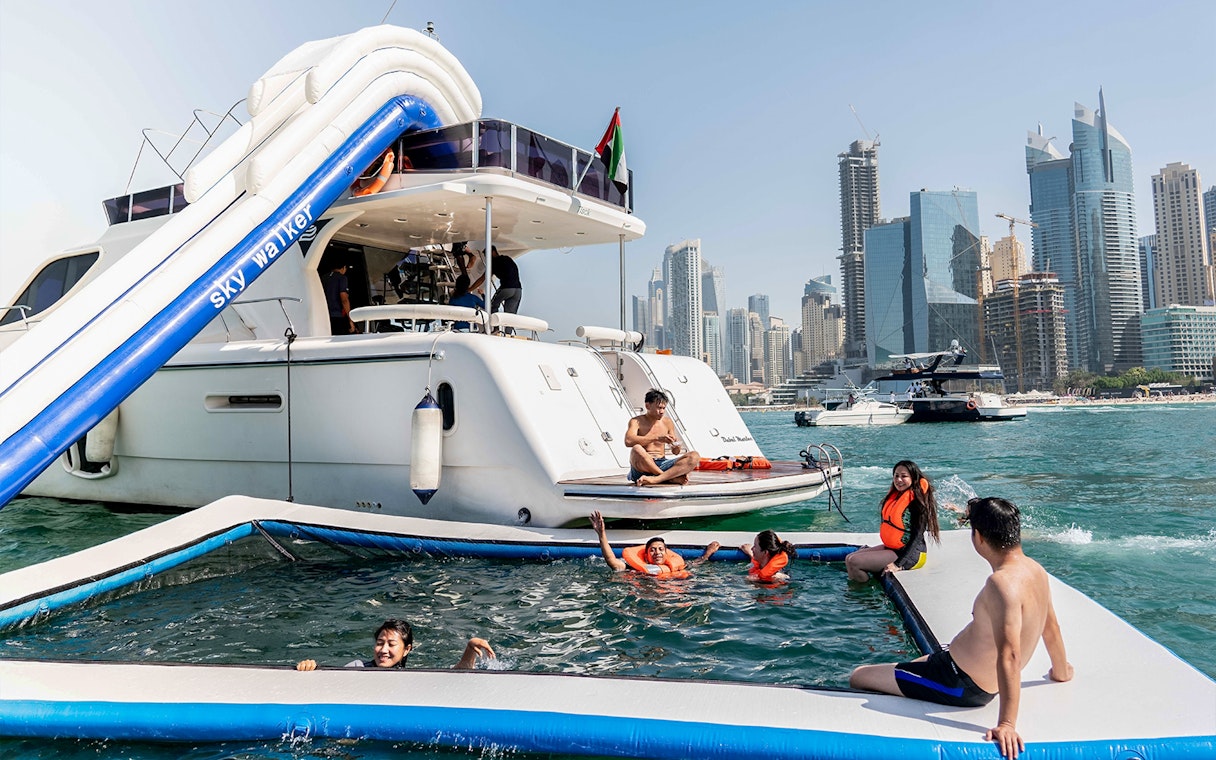 Yacht with slide and people swimming in Dubai Marina, skyscrapers in background.