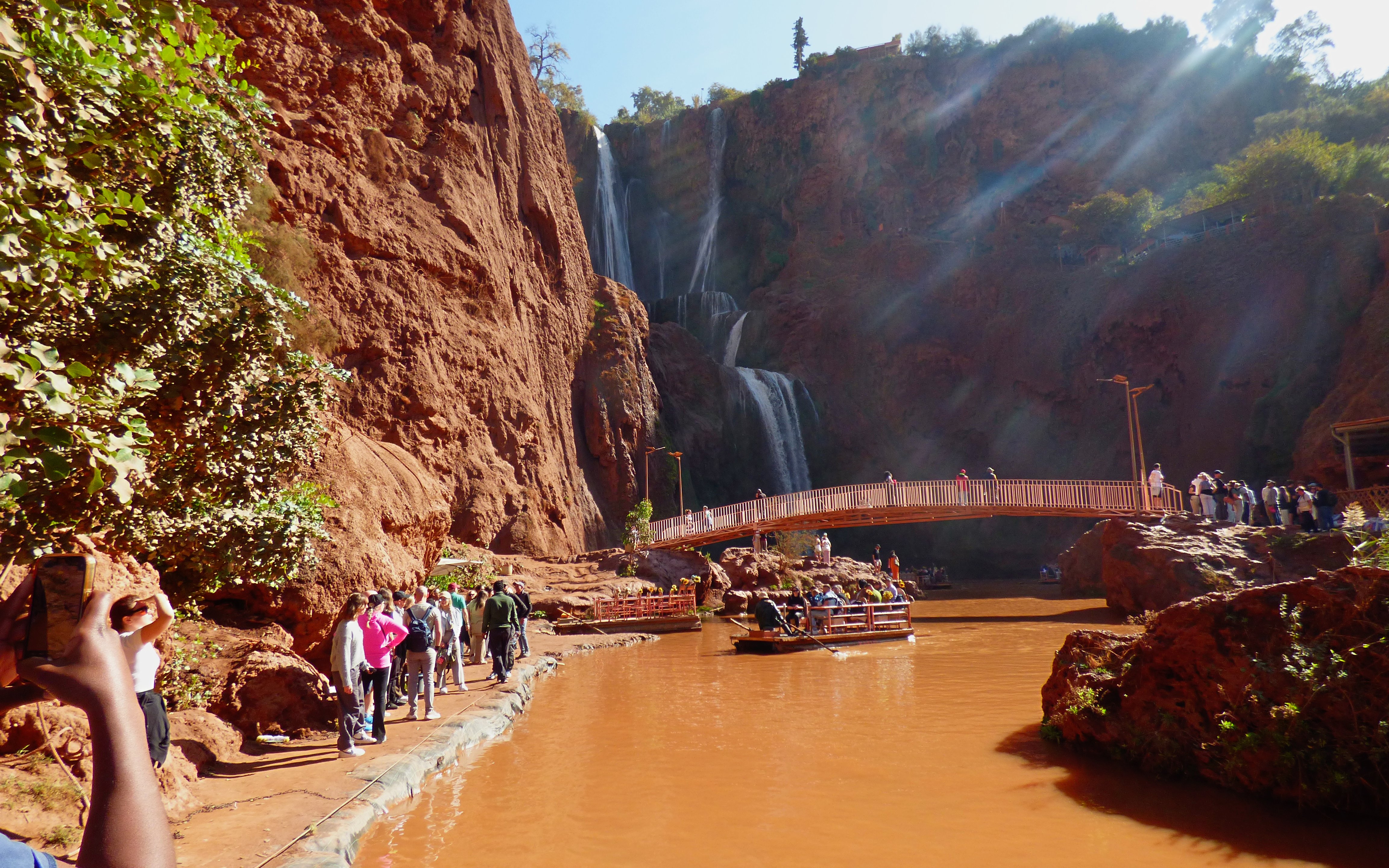 Tourists hiking near Ouzoud Waterfalls with a boat on the river and a bridge in the background.