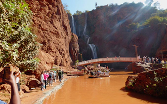 Tourists hiking near Ouzoud Waterfalls with a boat on the river and a bridge in the background.