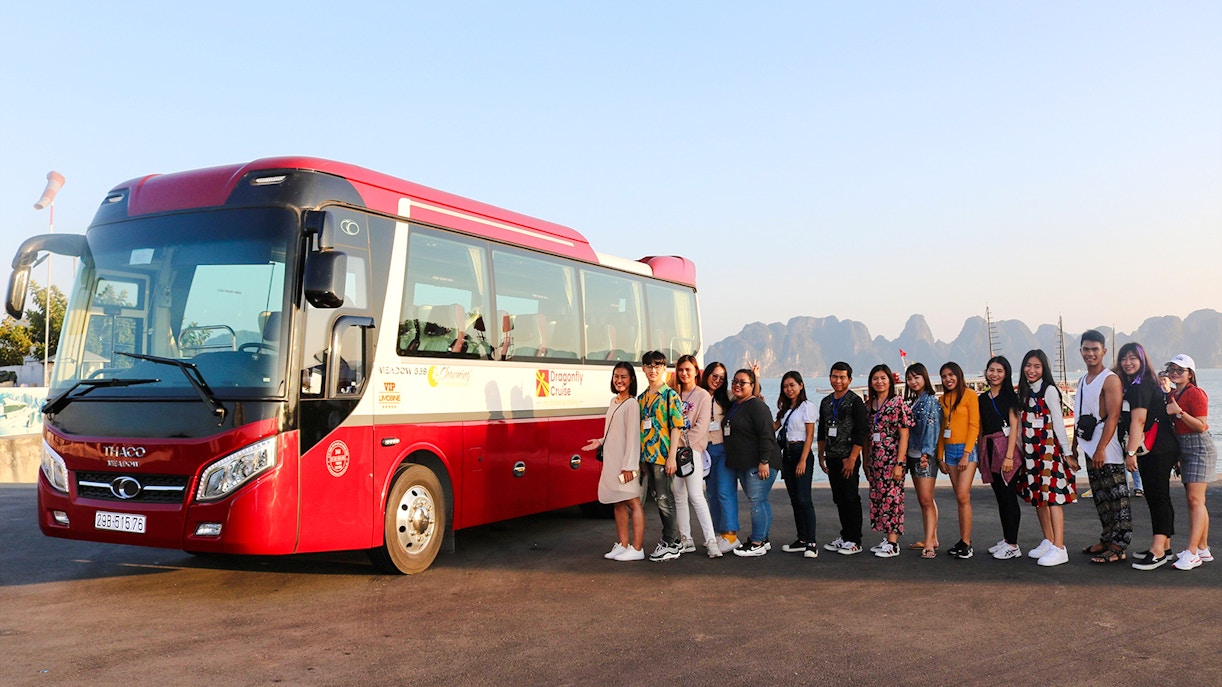 Travelers pose by a red tour bus with Ha Long Bay's karst formations in the background.