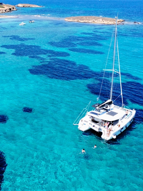 Catamaran sailing in clear turquoise waters near a rocky coastline.