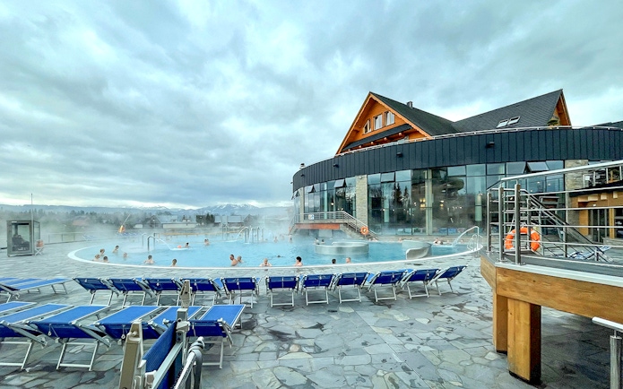 Outdoor pool at Zakopane Thermal Baths with people relaxing, surrounded by mountains.