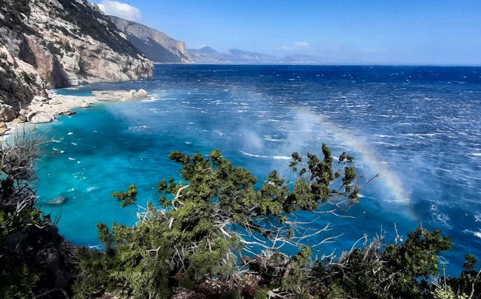 Coastal view of Cala Mariolu with clear blue water and a rainbow over the sea.