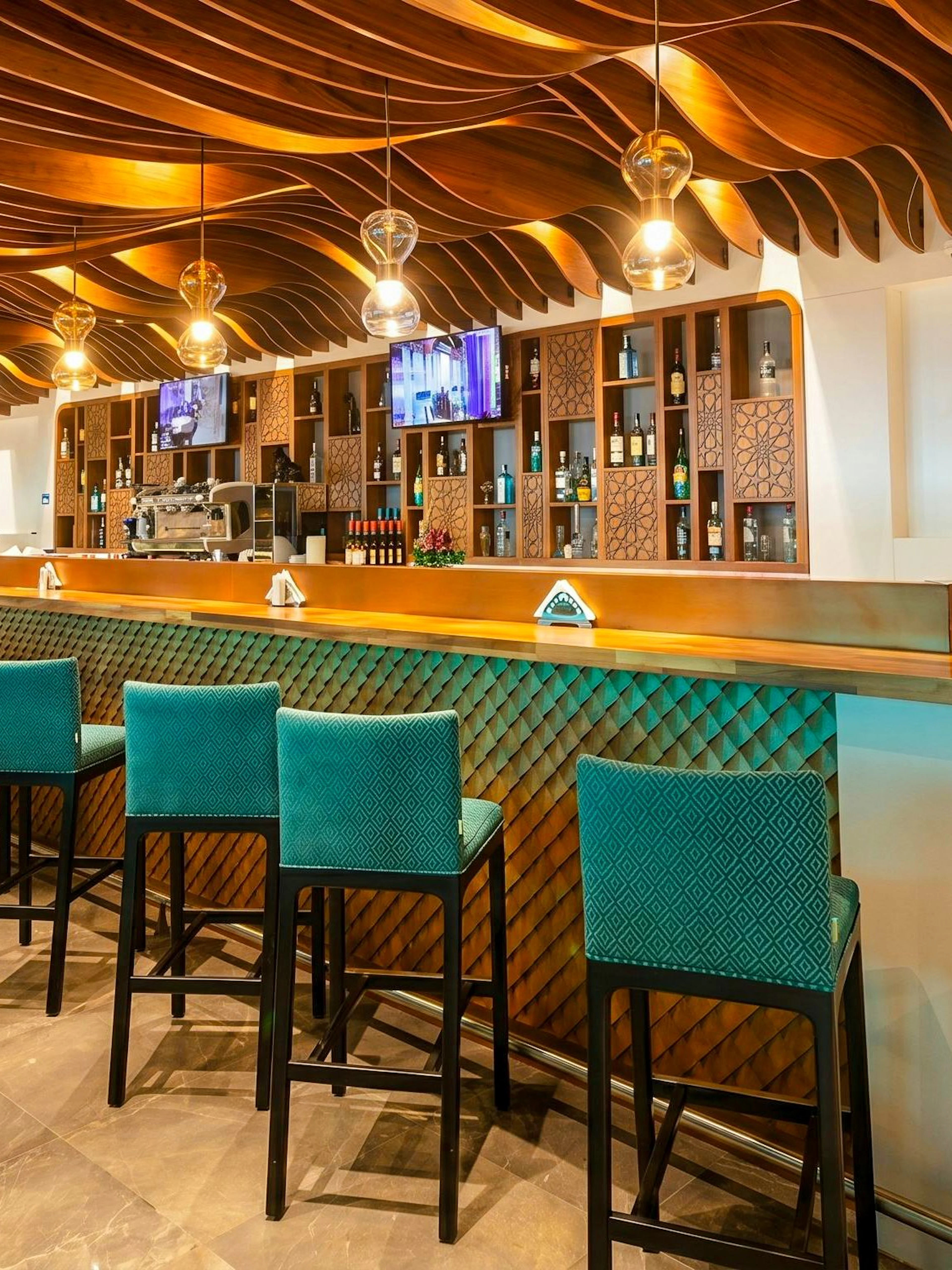 Bar area with teal chairs and wooden ceiling at Primeclass Lounge, Muscat International Airport.