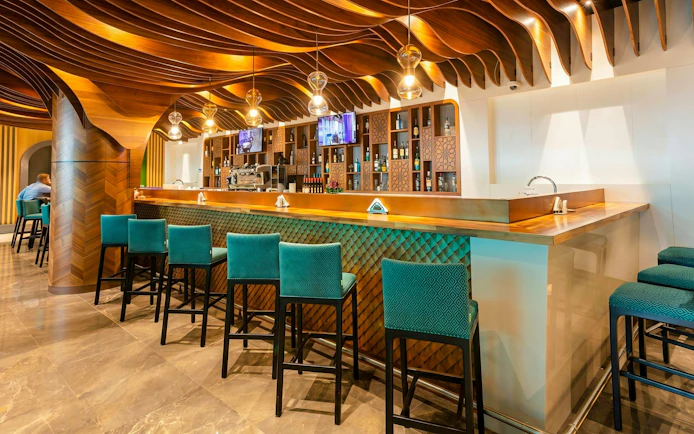 Bar area with teal chairs and wooden ceiling at Primeclass Lounge, Muscat International Airport.