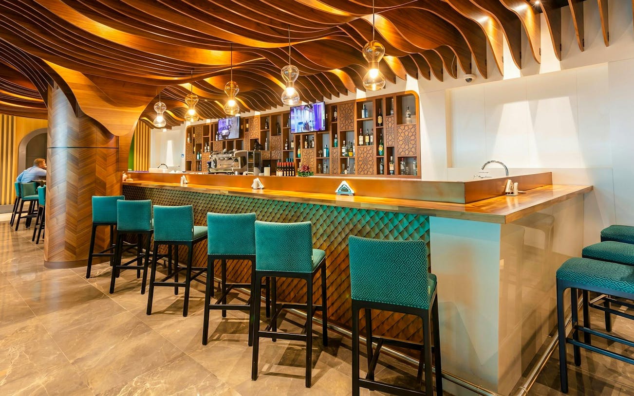 Bar area with teal chairs and wooden ceiling at Primeclass Lounge, Muscat International Airport.