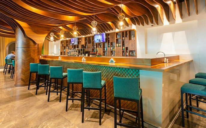 Bar area with teal chairs and wooden ceiling at Primeclass Lounge, Muscat International Airport.
