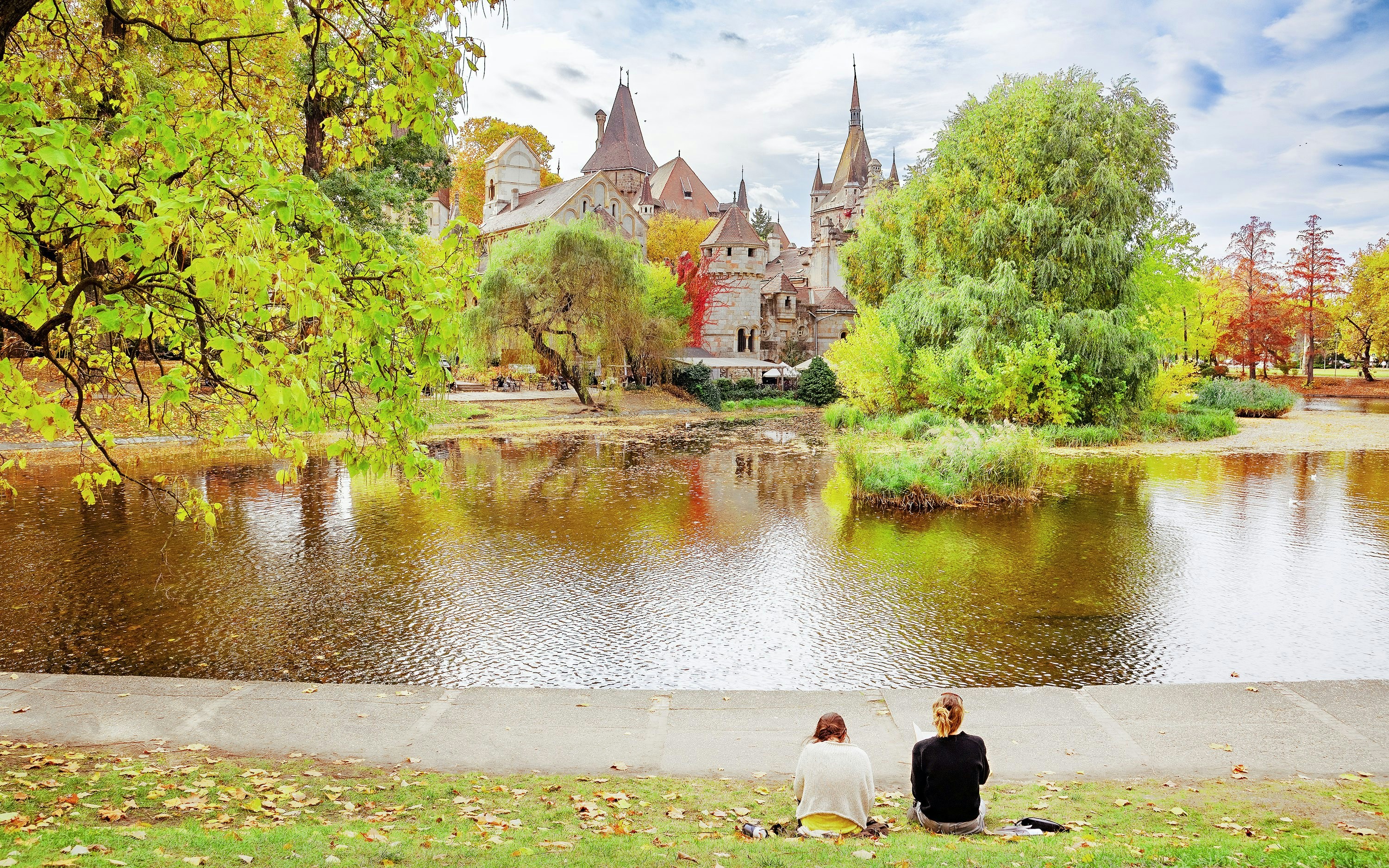 Two girls drawing by a lake in autumn, with Vajdahunyad Castle in the background.