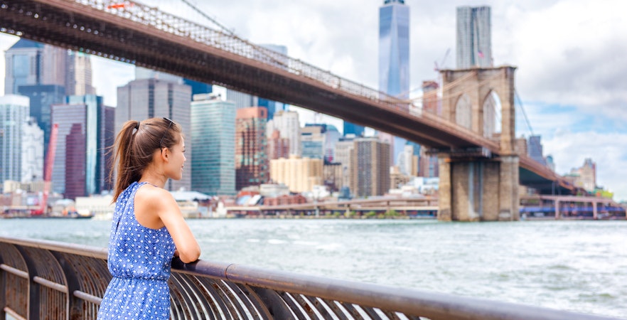 Woman in polka-dot dress views Lower Manhattan skyline from East River railing.