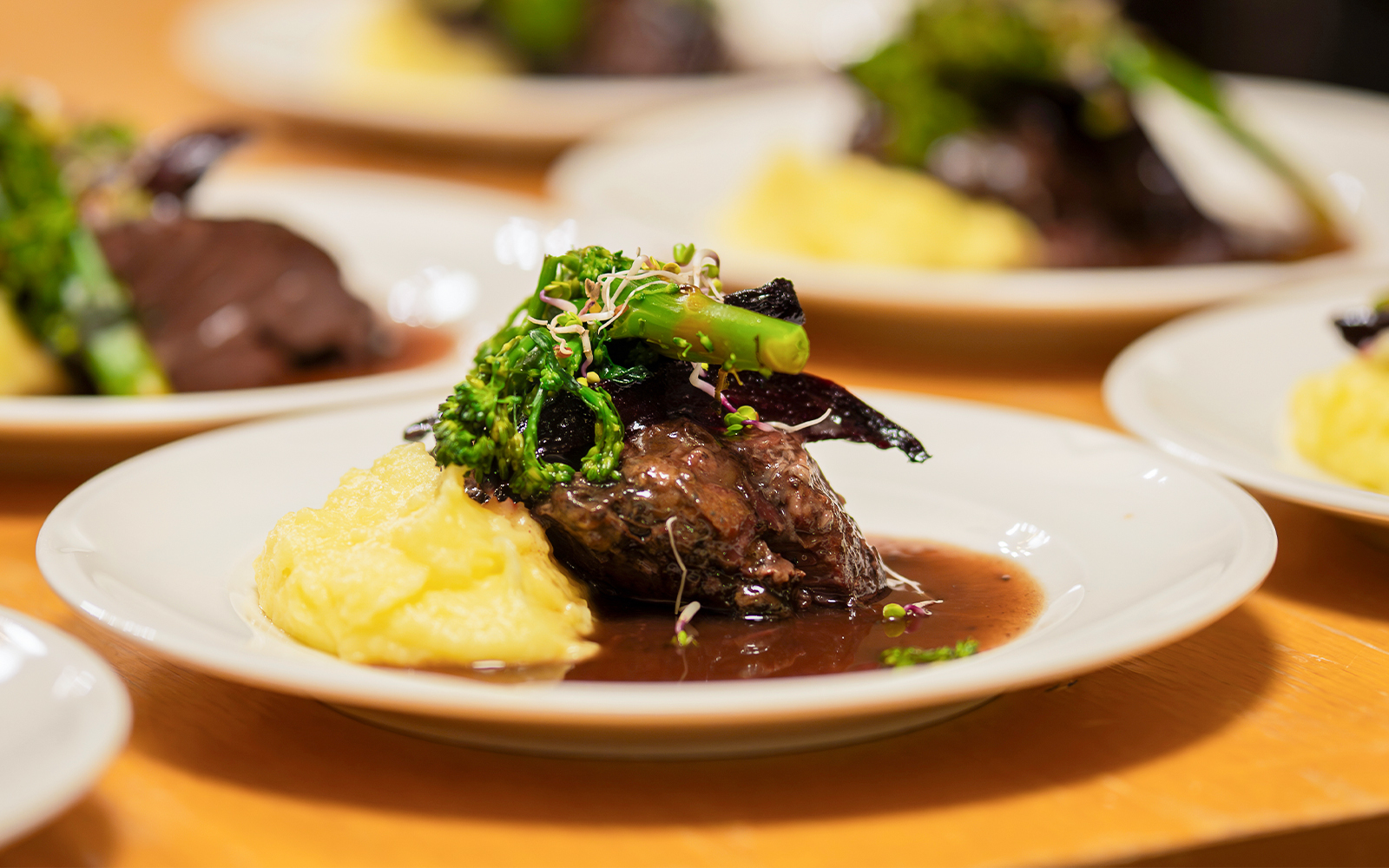 Slow cooked pork cheeks in red wine sauce with mashed potatoes and greens on a white plate.