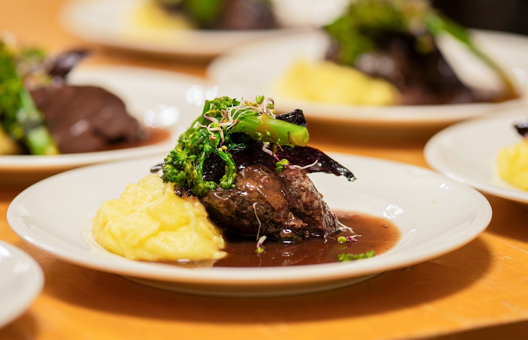 Slow cooked pork cheeks in red wine sauce with mashed potatoes and greens on a white plate.