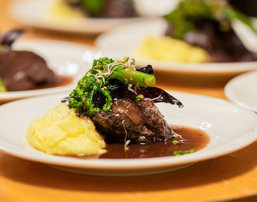 Slow cooked pork cheeks in red wine sauce with mashed potatoes and greens on a white plate.