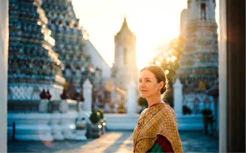 Tourist in traditional Thai costume at Wat Arun, Bangkok, with temple spires in the background.