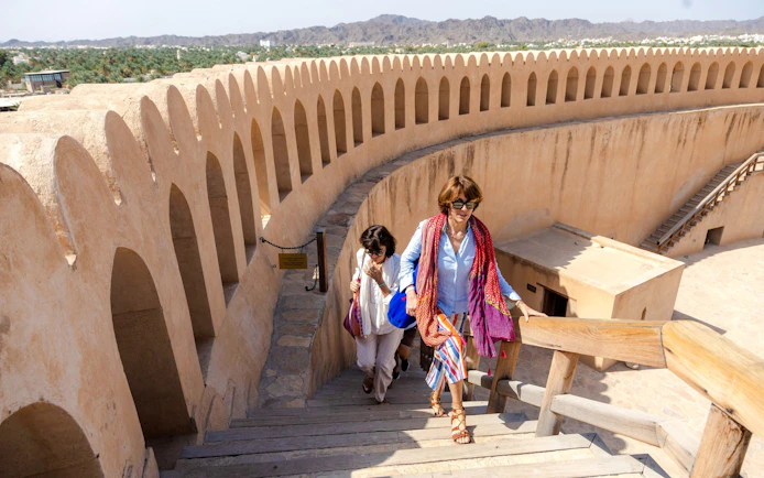 Visitors ascending stairs at Nizwa Fort, Oman, with mountain views in the background.