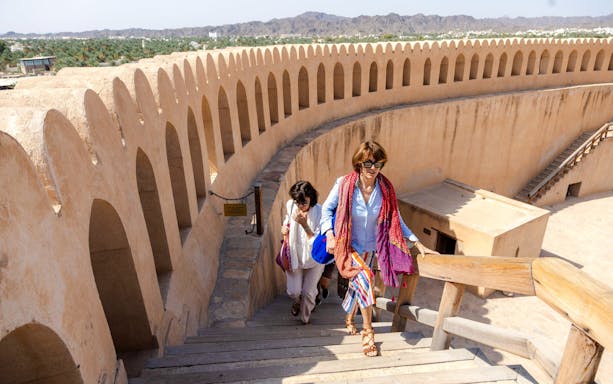 Visitors ascending stairs at Nizwa Fort, Oman, with mountain views in the background.