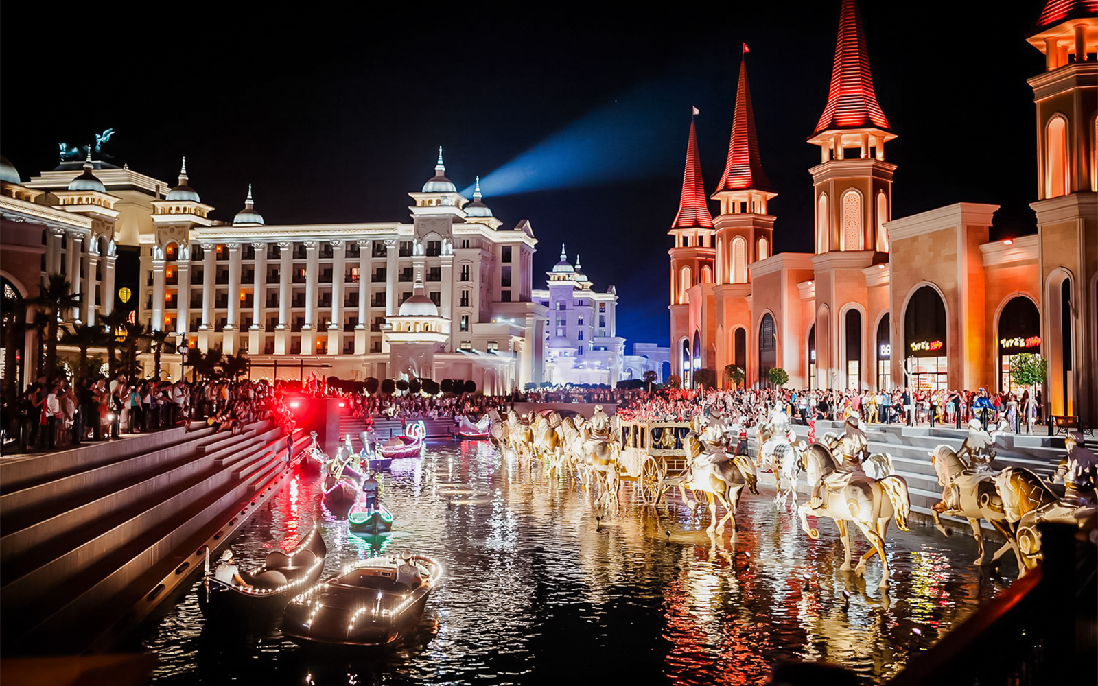 Boat parade at Land of Legends Night Show with illuminated buildings and crowd.