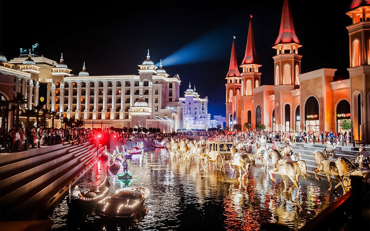 Boat parade at Land of Legends Night Show with illuminated buildings and crowd.