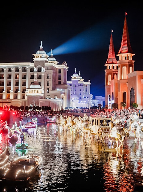 Boat parade at Land of Legends Night Show with illuminated buildings and crowd.