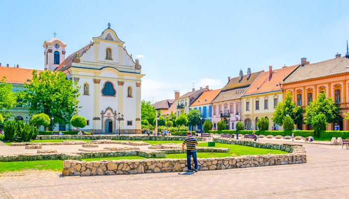 Feherek Church in Vic, Hungary, with colorful historic buildings and a stone courtyard.