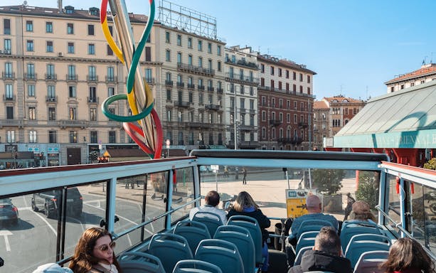 Open-top bus tour in Milan passing by colorful sculpture and historic buildings.