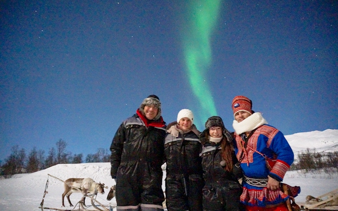 Group enjoying reindeer feeding under Northern Lights in snowy landscape.