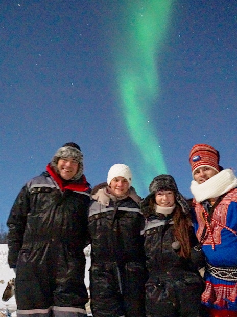 Group enjoying reindeer feeding under Northern Lights in snowy landscape.