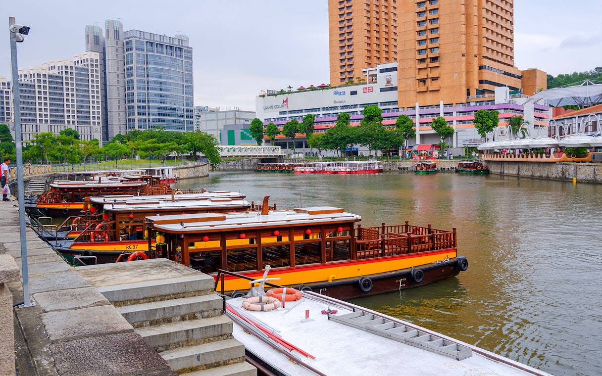 Traditional boats docked along the Singapore River with city buildings in the background.