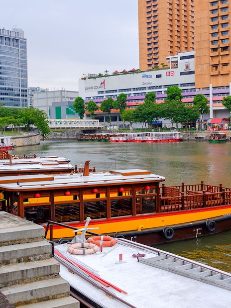 Traditional boats docked along the Singapore River with city buildings in the background.