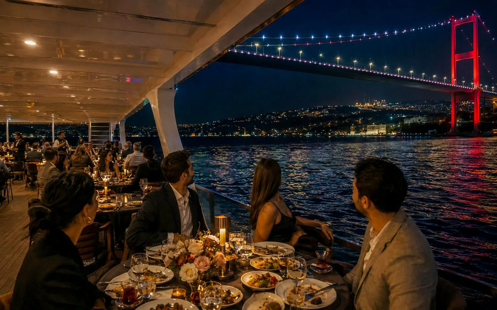 Guests dining on a yacht with a view of the illuminated Bosphorus Bridge at sunset, Istanbul.