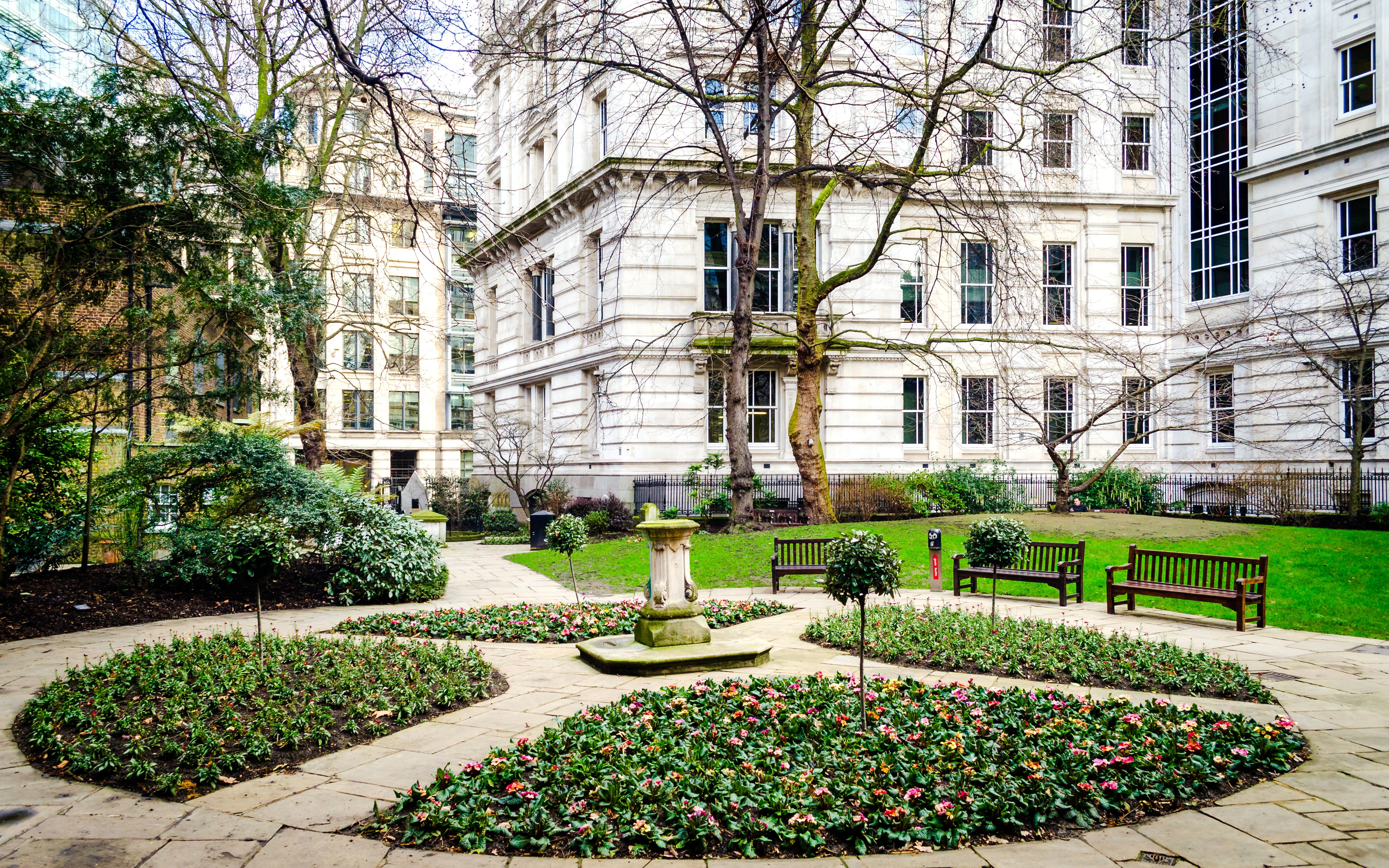 Postman's Park in London with flower beds, benches, and historic buildings in the background.