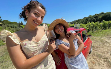Women enjoying wine by a red Fiat 500 in a Tuscan vineyard.