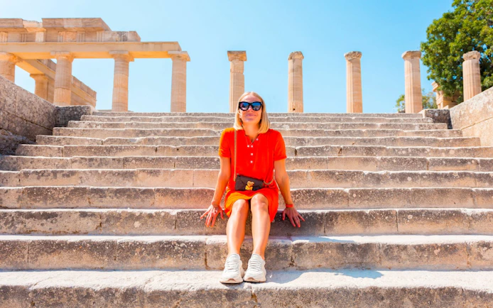 Visitor sitting on steps at Acropolis of Lindos, Greece, with ancient columns in background.