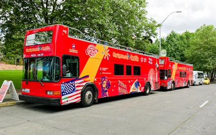Red double-decker tour bus in Seattle with sightseeing graphics parked by trees.