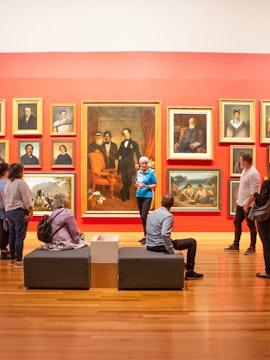 Group tour in an art gallery with visitors observing classic paintings on a red wall.
