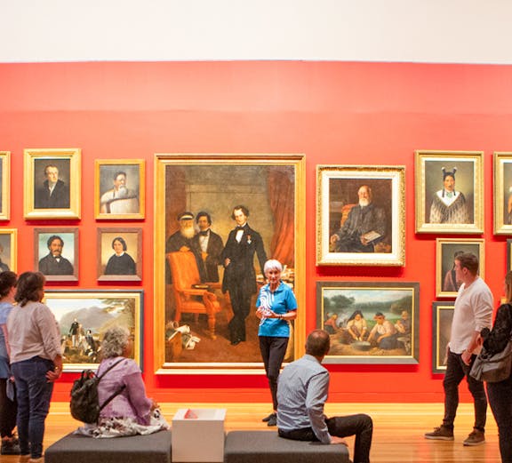 Group tour in an art gallery with visitors observing classic paintings on a red wall.
