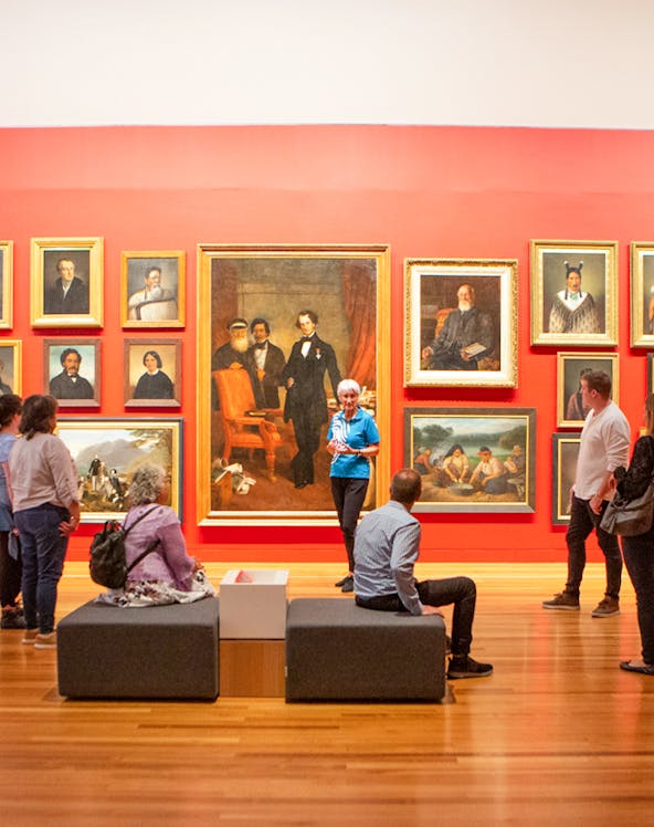 Group tour in an art gallery with visitors observing classic paintings on a red wall.