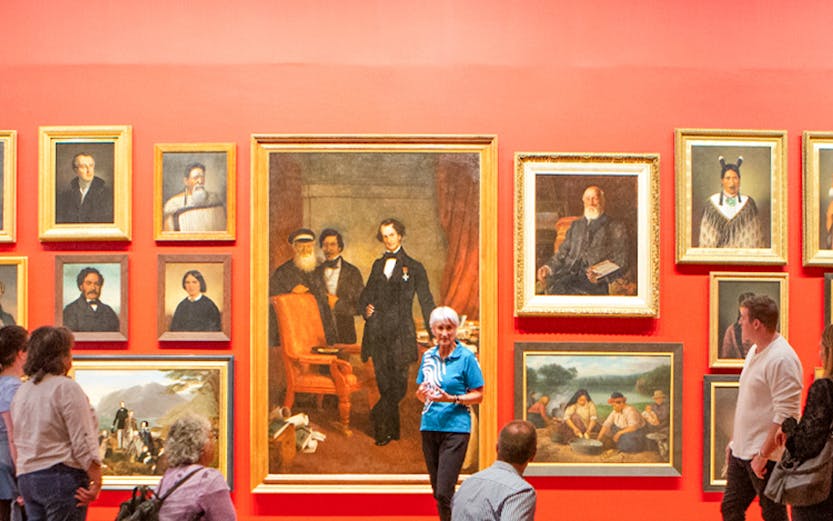 Group tour in an art gallery with visitors observing classic paintings on a red wall.