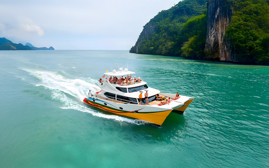 People enjoying a Langkawi sunset cruise on a catamaran near lush cliffs.