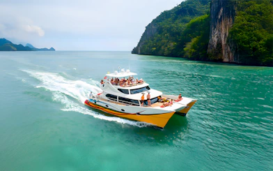 People enjoying a Langkawi sunset cruise on a catamaran near lush cliffs.