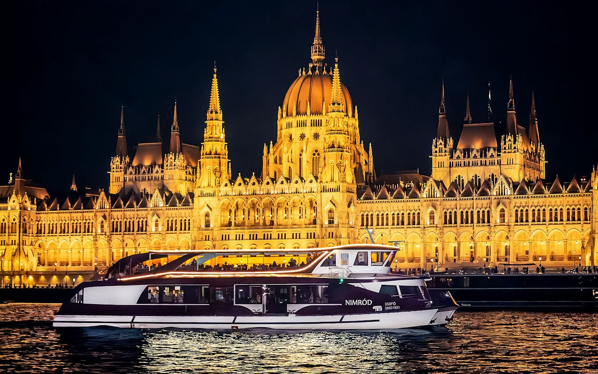 Cruise boat on Danube River at night with Budapest Parliament illuminated in background.