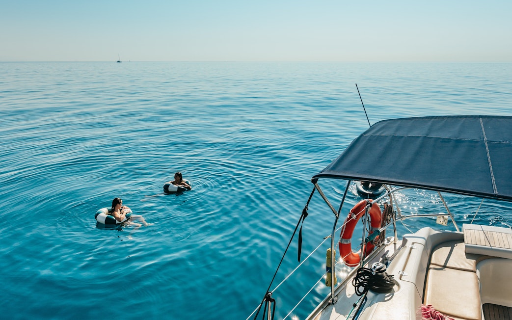 Friends swimming near a sailing yacht on a Barcelona sightseeing cruise.