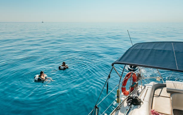 Friends swimming near a sailing yacht on a Barcelona sightseeing cruise.
