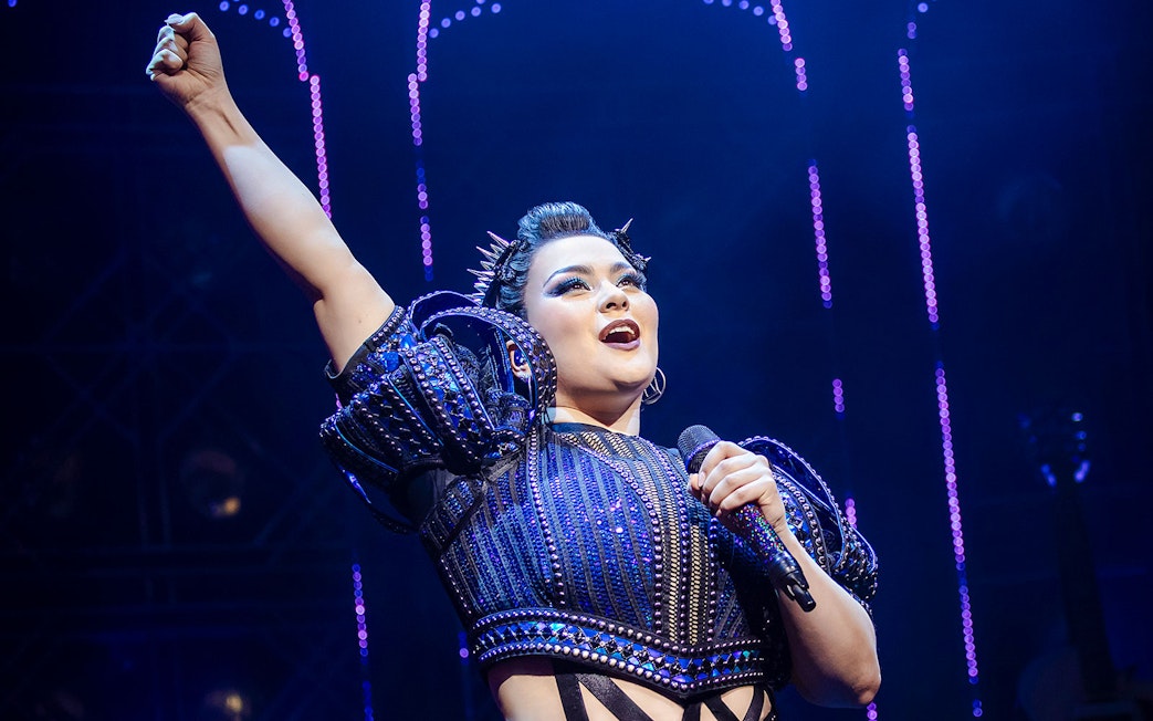 Performer on stage during the musical "Six" with microphone and raised arm.