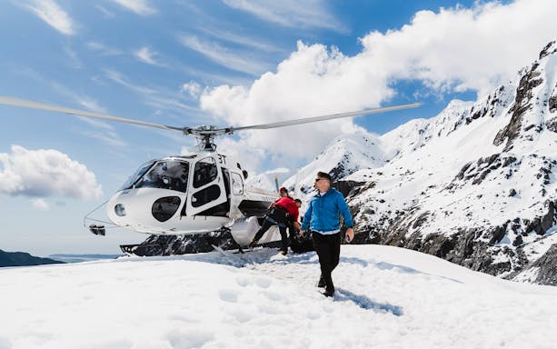 Helicopter landing on snowy glacier with passengers disembarking, surrounded by mountains.