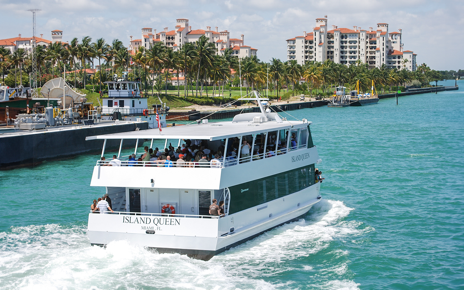 Sightseeing cruise boat in Miami waters with waterfront buildings and palm trees in the background.