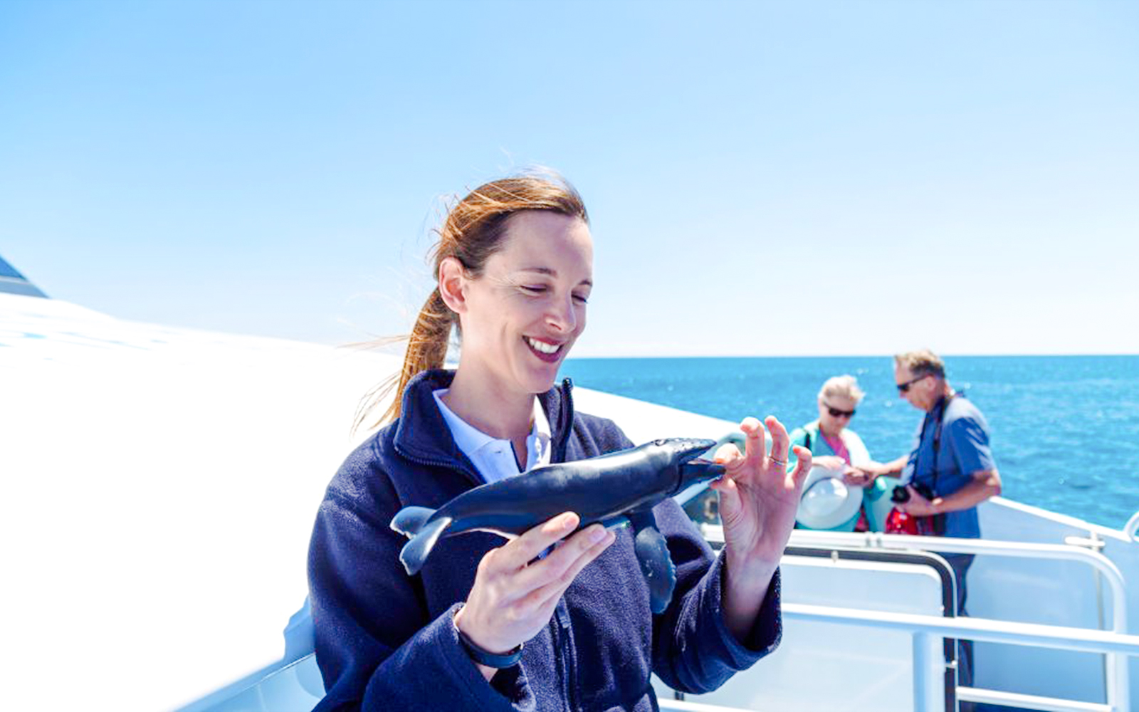 Guide holding model whale on boat with tourists in background.